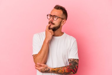 Pembe arka planda izole edilmiş, şüpheli ve şüpheci bir ifadeyle yan bakan beyaz dövmeli genç bir adam..