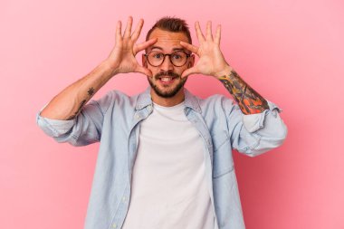 Pembe arka planda izole edilmiş dövmeleri olan beyaz genç bir adam bir başarı fırsatı bulmak için gözlerini açık tutuyor..