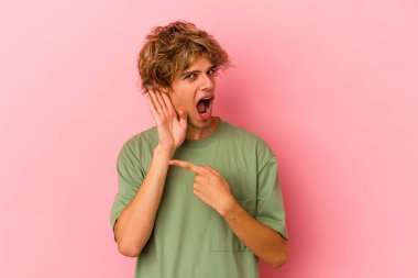 Pembe arka planda soyutlanmış beyaz makyajlı genç bir adam bir dedikoduyu dinlemeye çalışıyor..