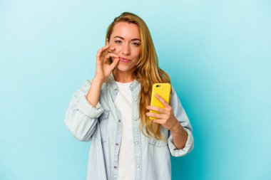 Cep telefonunu elinde tutan beyaz kadın mavi arka planda izole edilmiş ve parmakları dudaklarında bir sır saklıyor..