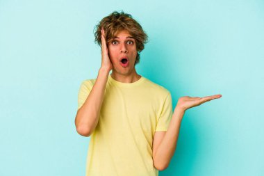 Mavi arka planda izole edilmiş beyaz makyajlı genç bir adam avuç içi fotokopi alanını elinde tutmaktan etkilenmiş..