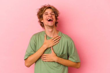 Pembe arka planda soyutlanmış beyaz makyajlı genç bir adam yüksek sesle gülüyor ve elini göğsüne koyuyor..