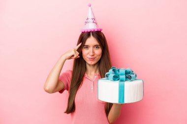 Beyaz bir kadın pembe arka planda izole edilmiş bir doğum gününü kutluyor. Şakağı işaret ediyor, düşünüyor, bir göreve odaklanıyor..