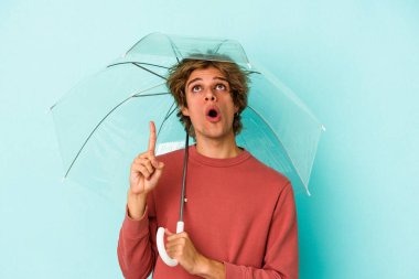 Makyaj şemsiyesi tutan genç beyaz adam mavi arka planda tek başına ağzı açık, üst tarafı işaret ediyor..