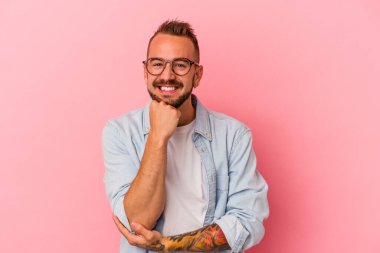 Pembe arka planda izole edilmiş, mutlu ve kendinden emin gülümseyen, elleriyle çenesine dokunan beyaz dövmeli genç bir adam..