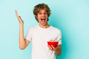 Makyajlı genç beyaz adam mavi arka planda izole edilmiş bir mısır gevreği kasesi hoş bir sürpriz alıyor, heyecanlı ve el kaldırıyor..