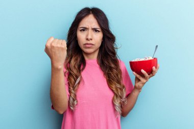 Mavi arka planda izole edilmiş mısır gevreği kasesi tutan genç bir ırk kadını kameraya yumruk gösteriyor, agresif bir yüz ifadesi..