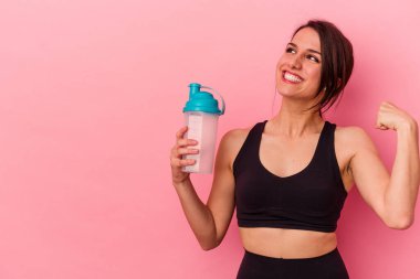 Pembe arka planda izole edilmiş protein içeceği içen beyaz bir kadın zaferden sonra yumruğunu kaldırıyor..