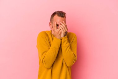 Pembe arka planda izole edilmiş dövmeleri olan beyaz genç bir adam parmaklarıyla kameraya göz kırpıyor, yüzünü kapatmaktan utanıyor..