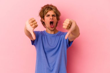 Pembe arka planda soyutlanmış beyaz tenli genç bir adam baş parmağını aşağı gösteriyor ve hoşlanmadığını ifade ediyor..