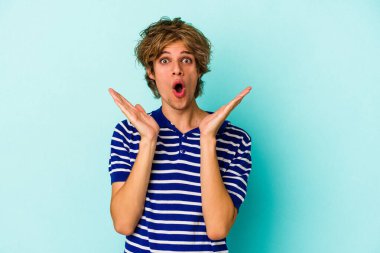 Makyajlı genç beyaz adam mavi arka planda izole edilmiş, şaşırmış ve şok olmuş..