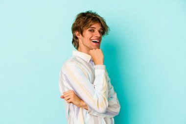 Mavi arka planda soyutlanmış, mutlu ve kendinden emin gülümseyen, elleriyle çenesine dokunan beyaz bir genç..