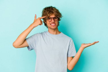 Makyajlı genç beyaz adam mavi arka planda izole edilmiş ve elinde bir ürün var..