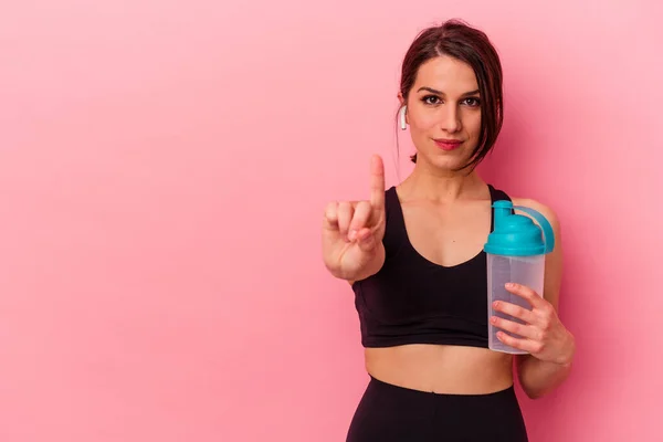 ピンクの背景に単離されたタンパク質シェイクを飲む若い白人女性は指でナンバーワンを示す — ストック写真