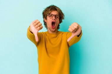 Mavi arka planda soyutlanmış beyaz tenli genç bir adam baş parmağını aşağı gösteriyor ve hoşlanmadığını ifade ediyor..