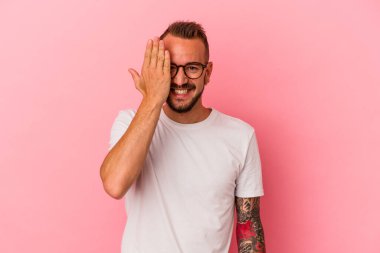 Pembe arka planda dövmeleri olan beyaz bir adam yüzünün yarısını avuç içiyle örterek eğleniyor..