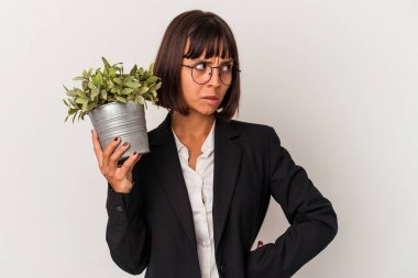 Genç melez iş kadını beyaz arka planda izole edilmiş bir bitki tutuyor. Kafası karışmış, şüpheli ve güvensiz hissediyor..