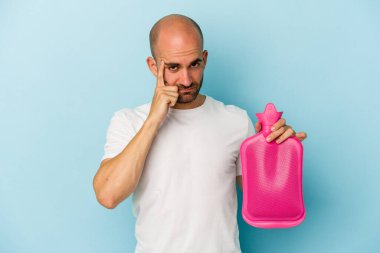 Mavi arka planda sıcak bir su torbası tutan genç kel bir adam şakağı parmağıyla işaret ediyor, düşünüyor ve bir göreve odaklanıyor..