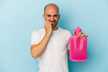 Mavi arka planda izole edilmiş sıcak bir su torbası tutan genç kel bir adam tırnaklarını yiyor, gergin ve çok endişeli..