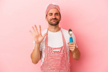 Genç dondurmacı elinde kaşık tutan pembe arka planda izole edilmiş gülümseyen beş numarayı parmaklarıyla gösteriyor..