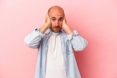 Kafkasyalı genç kel adam pembe arka planda izole edilmiş kulakları kulaklarıyla kapatıyor çok fazla ses duymamaya çalışıyor..