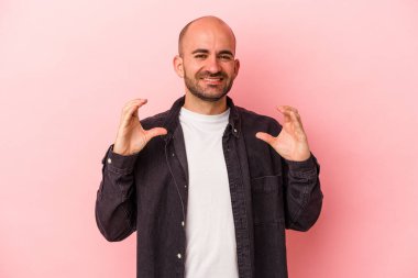 Beyaz, kel bir adam pembe arka planda izole edilmiş. Elinde avuç içi olan bir şeyle kameraya sunuyor..