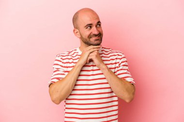 Pembe arka planda izole edilmiş genç beyaz kel adam ellerini çenesinin altında tutuyor, mutlu bir şekilde yana bakıyor..