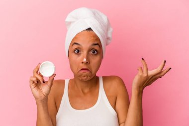 Yüzüne krem süren genç melez kadın pembe arka planda izole edilmiş omuzlar ve açık gözler..