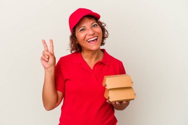 Beyaz arka planda izole edilmiş burgerleri olan orta yaşlı bir teslimatçı kadın neşeli ve kaygısız parmaklarıyla barış sembolü gösteriyor..