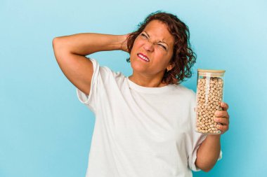 Orta yaşlı Latin bir kadın elinde nohut kavanozu tutarken mavi arka planda izole edilmiş kafasının arkasına dokunuyor, düşünüyor ve bir seçim yapıyor..