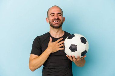Genç iş adamı kel adam mavi arka planda tek başına futbol oynuyor kahkahalar atıyor elini göğsüne koyuyor.