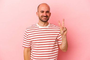 Genç beyaz kel adam pembe arka planda izole edilmiş neşeli ve kaygısız parmaklarıyla barış sembolü gösteriyor..