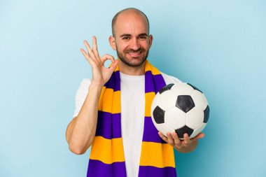 Mavi arka planda izole edilmiş atkı takan genç ve kel bir adam neşeli ve kendinden emin..