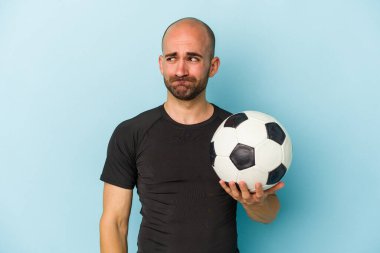 Genç iş adamı kel adam mavi arka planda tek başına futbol oynuyor. Kafası karışmış, şüpheli ve kendinden emin değil..