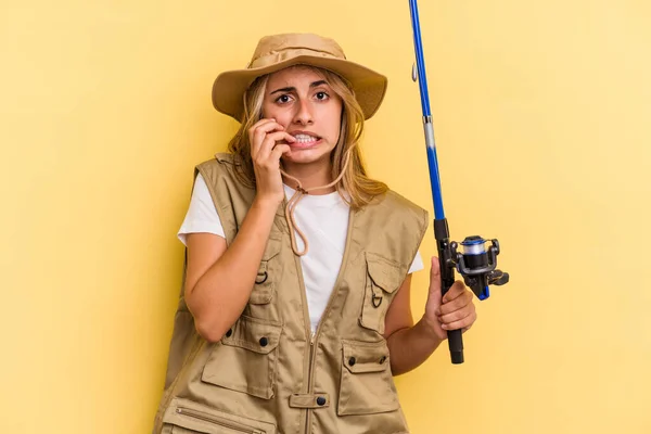 年轻的高加索金发女渔夫拿着一根用黄色背景隔离的钓竿 咬着指甲 紧张而焦虑 — 图库照片