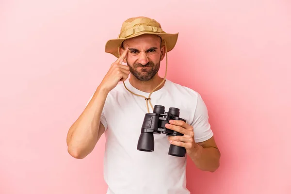 年轻的高加索秃头男子手持双筒望远镜 呈粉红色背景 用食指表示失望的姿态 — 图库照片