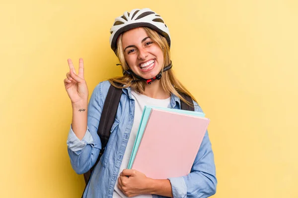 黄色の背景に隔離された自転車のヘルメットを身に着けている若い白人学生の女性喜びと屈託のない指で平和のシンボルを示す — ストック写真