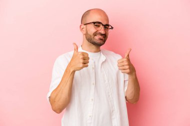 Genç beyaz kel adam pembe arka planda izole edilmiş. Baş parmağını kaldırıyor, gülümsüyor ve kendinden emin..