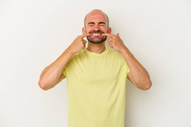 Beyaz arka planda soyutlanmış genç kel adam gülümsüyor, parmaklarını ağıza doğrultuyor..