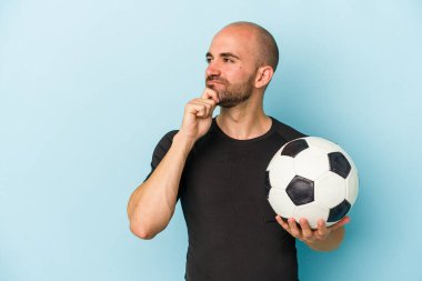 Genç iş adamı kel adam mavi arka planda izole bir şekilde futbol oynuyor. Şüpheli ve şüpheci bir ifadeyle yana bakıyor..