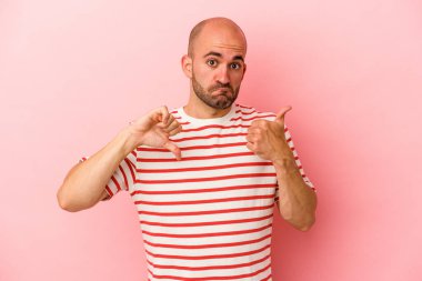 Pembe arka planda izole edilmiş genç, beyaz, kel bir adam. Baş parmağını kaldırıyor, baş parmaklarını eğiyor. Zor bir seçim.