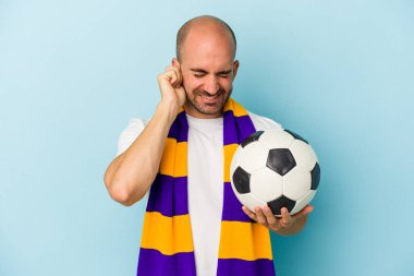 Mavi arka planda izole edilmiş eşarp takan kel bir genç adam kulaklarını elleriyle kapatıyor..