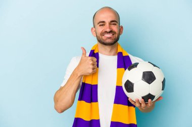 Mavi arka planda izole edilmiş atkı takan genç ve kel bir adam gülümsüyor ve başparmağını kaldırıyor.