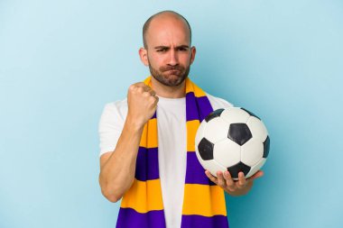 Mavi arka planda izole edilmiş atkı takan genç ve kel bir adam kameraya yumruğunu gösteriyor, agresif bir yüz ifadesi..