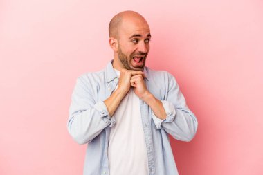 Pembe arka planda izole edilmiş genç beyaz kel adam ellerini çenesinin altında tutuyor, mutlu bir şekilde yana bakıyor..