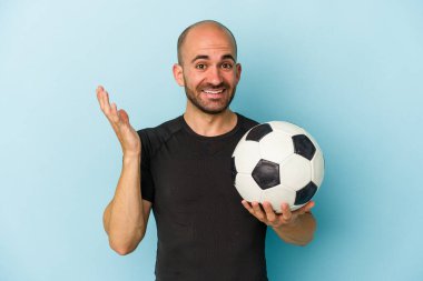 Genç iş adamı kel adam mavi arka planda izole bir şekilde futbol oynuyor. Hoş bir sürpriz, heyecanlı ve el kaldırıyor..