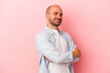 Pembe arka planda kendini güvende hisseden, kararlılıkla kollarını kavuşturan beyaz kel genç bir adam..