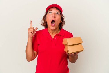 Beyaz arka planda soyulmuş burgerleri alan orta yaşlı teslimatçı kadın ağzı açık bir şekilde üst tarafı işaret ediyor..