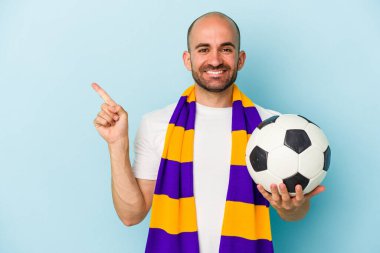 Mavi arka planda izole edilmiş atkı takan genç ve kel bir adam gülümsüyor ve boş alanda bir şeyler gösteriyor..