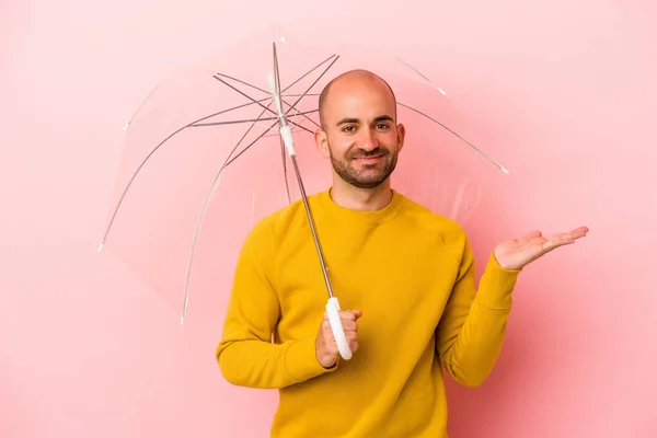 ピンクの背景に隔離された傘を持っている若い白人のはげ男は 手のひらにコピースペースを示し 腰に別の手を保持します — ストック写真